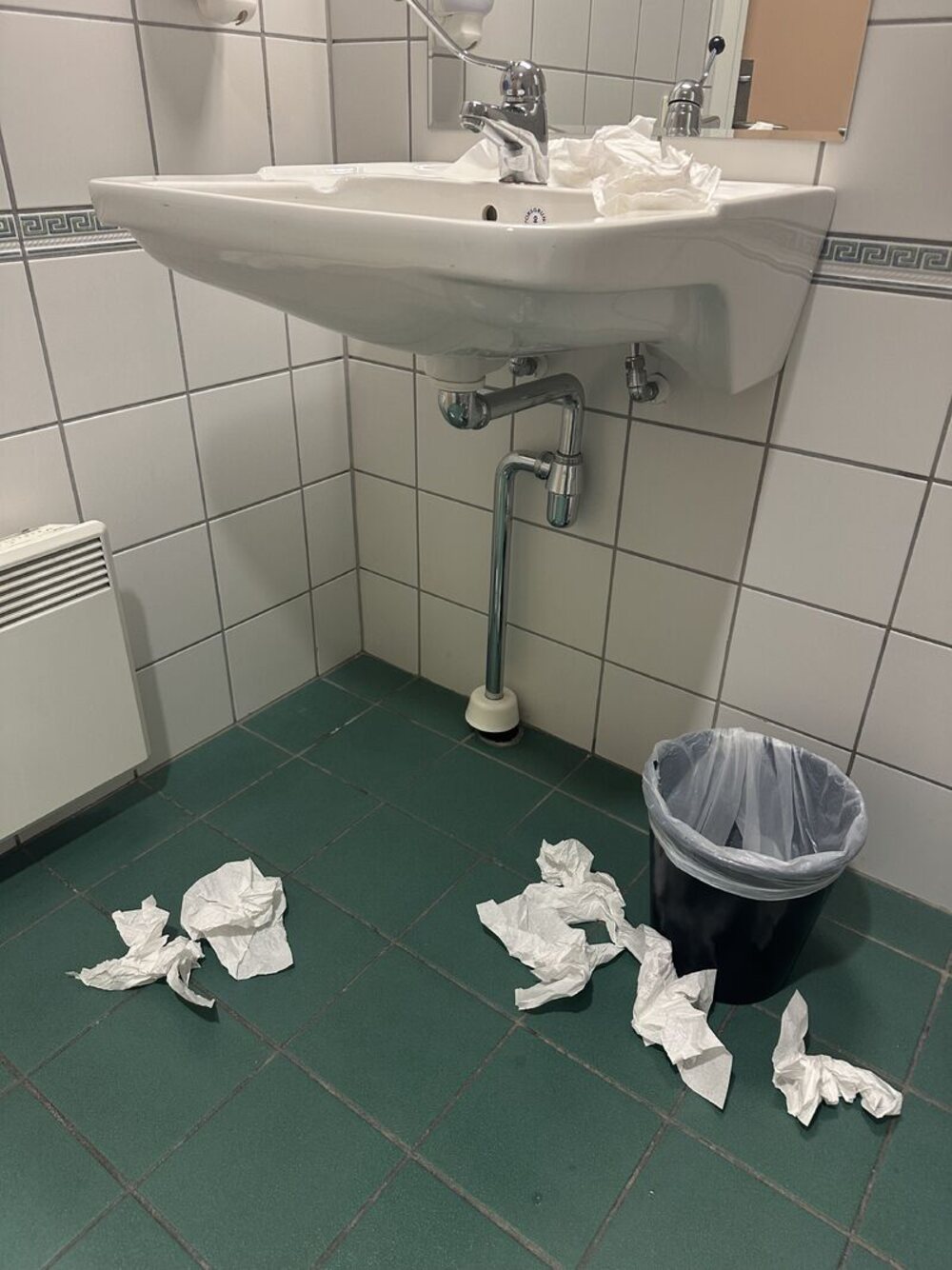 Et toalettrom der det ligger tørkepapir på gulvet rundt vasken og søppelbøtta